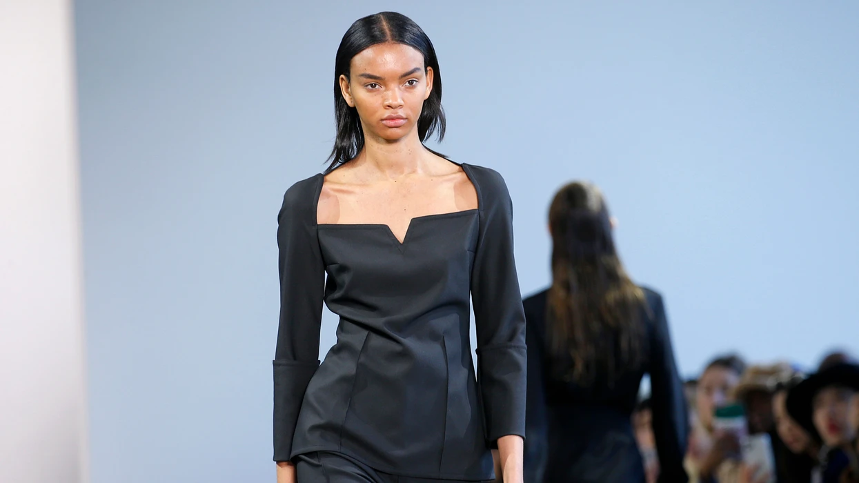 A model wearing a simple, elegant black outfit walks down the runway, embodying minimalist fashion.